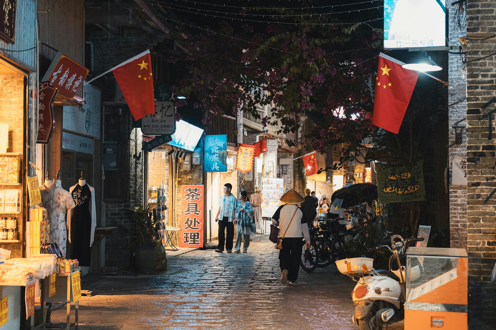 People walk down a narrow street with chinese flags.